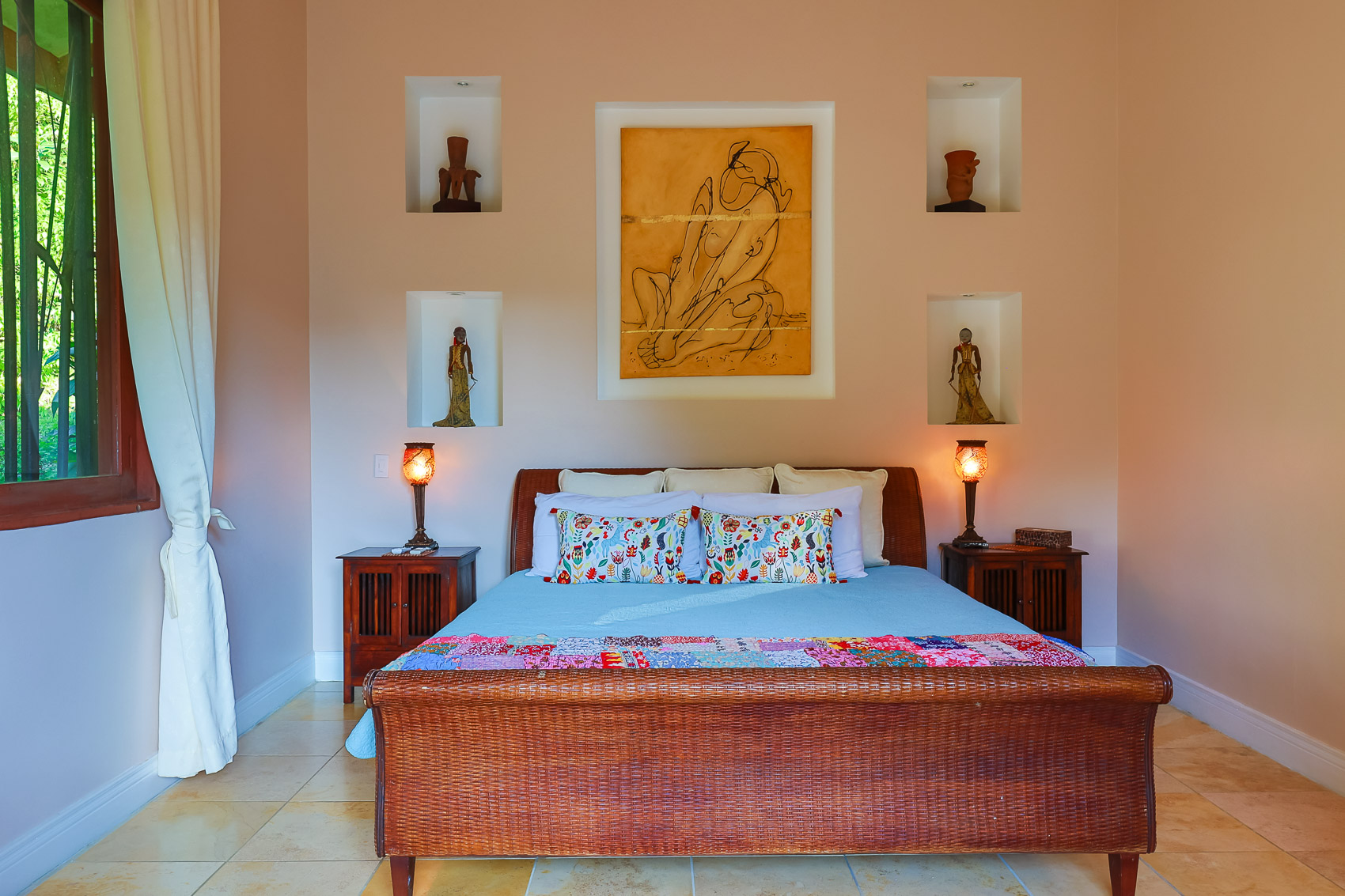 A bedroom features a woven bed frame, built-in wall niches with artwork, and a window providing natural light and a view of lush greenery.