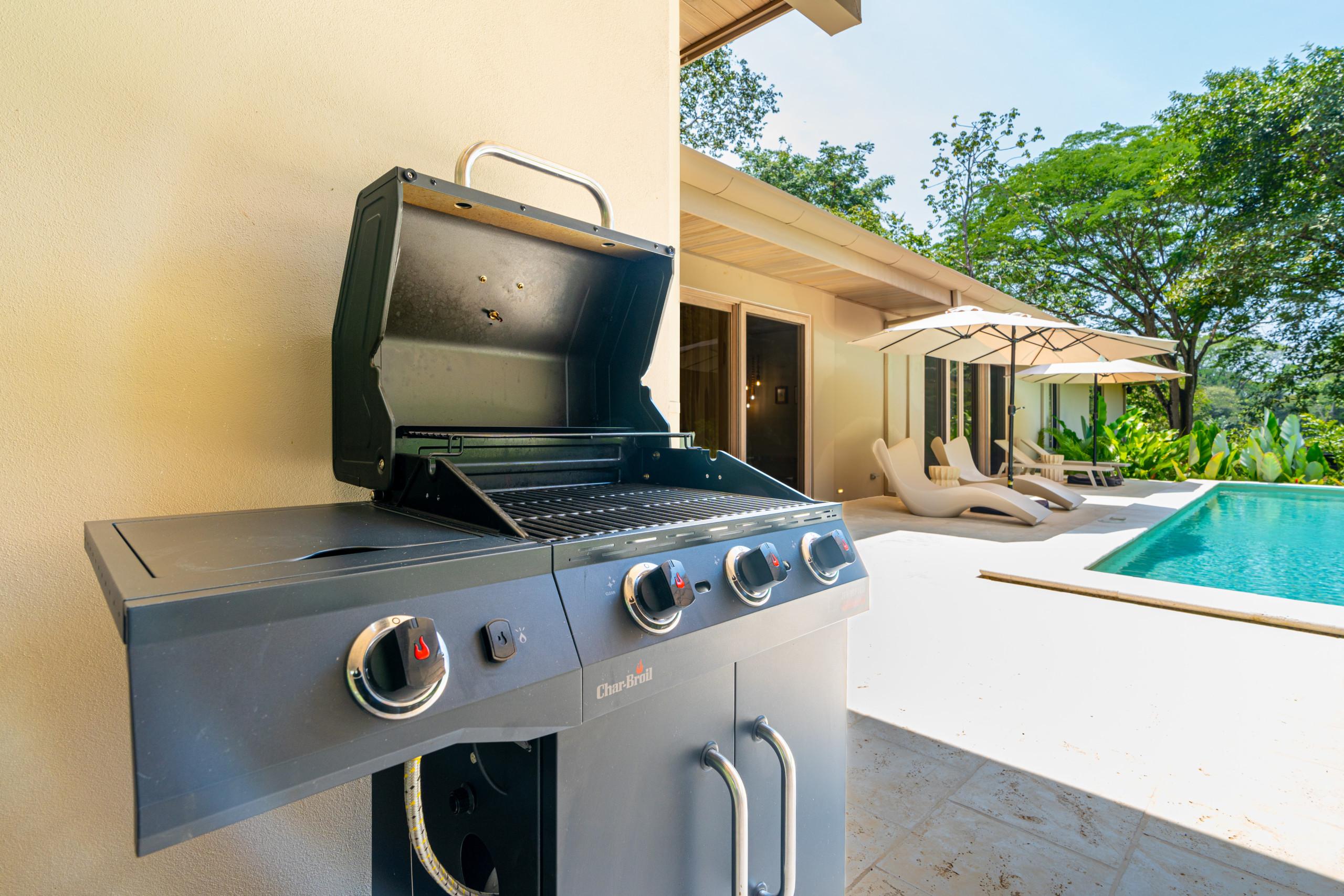 The sunny outdoor patio features a gas grill, leading to a private pool area with sun loungers and umbrellas, all set against a backdrop of lush jungle.