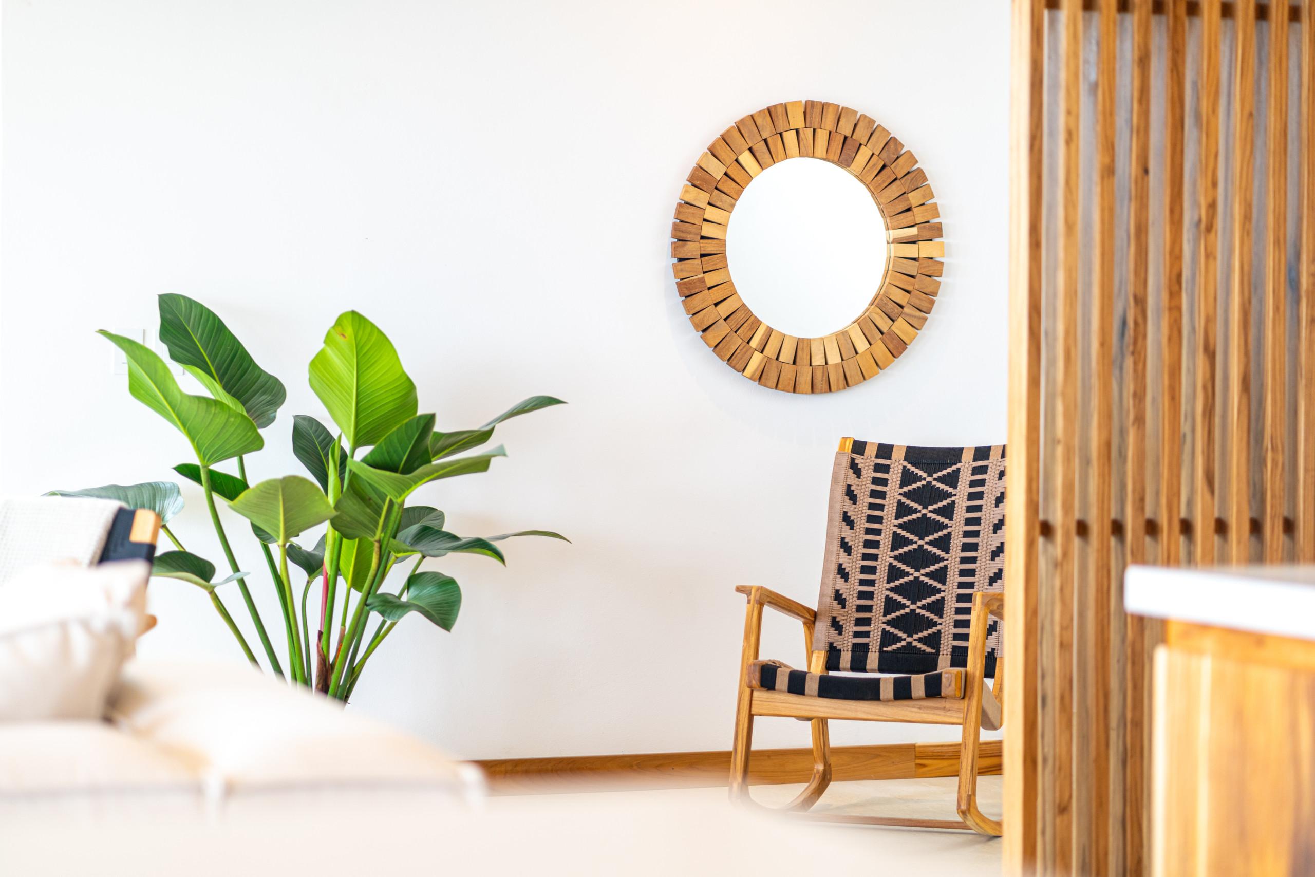 A bright interior space features a Masaya rocking chair, a large tropical plant, and a decorative mirror set against a white wall and a natural wood slatted partition.