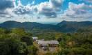 An aerial view reveals the villa's structures nestled among lush tropical jungle, extending to distant mountains and the Pacific Ocean horizon.