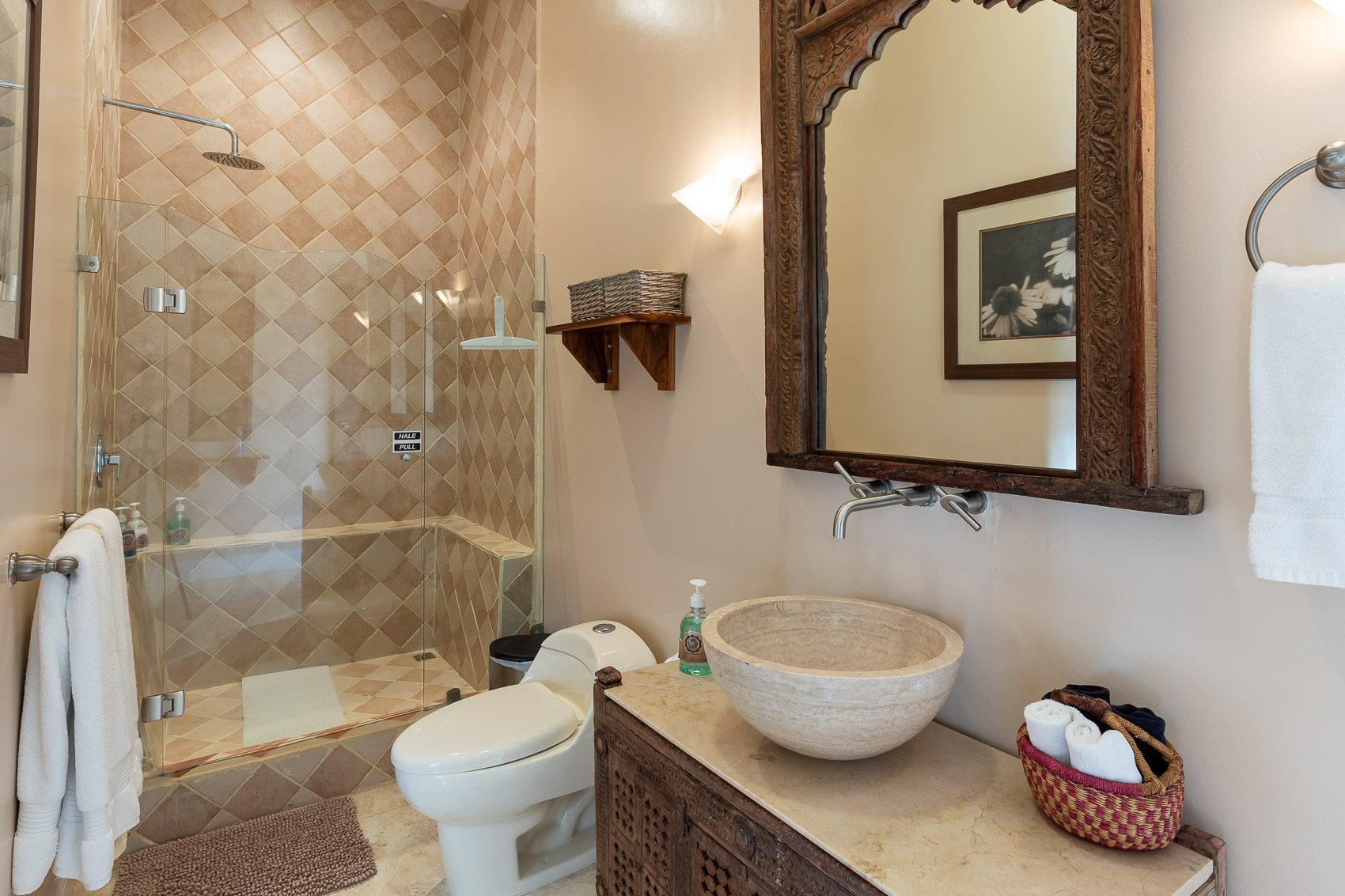 A Balinese-inspired bathroom offers a walk-in shower with warm-toned diamond-patterned tiles, a stone vessel sink on a vanity, and an intricately carved wooden mirror.