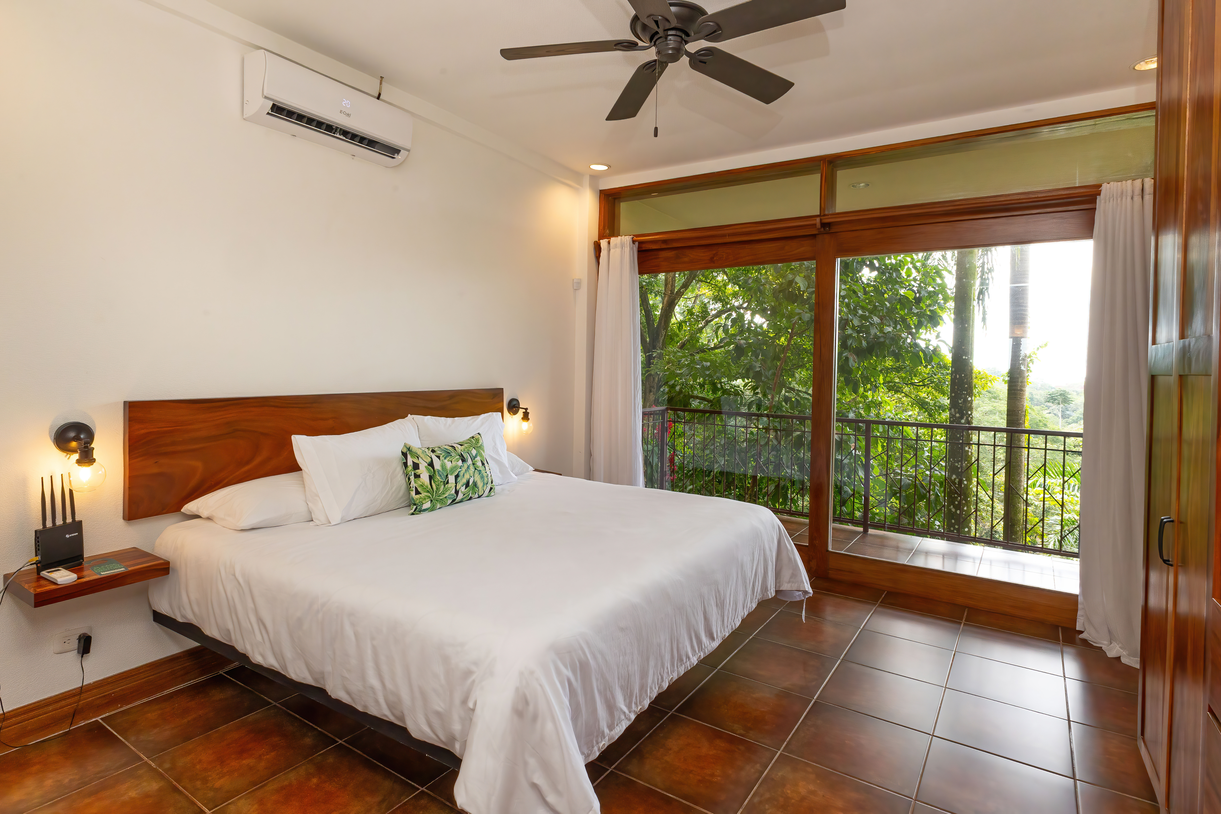 A serene king bedroom features a ceiling fan and air conditioning, with a large wooden-framed sliding glass door providing natural light and access to a private balcony overlooking the rainforest.