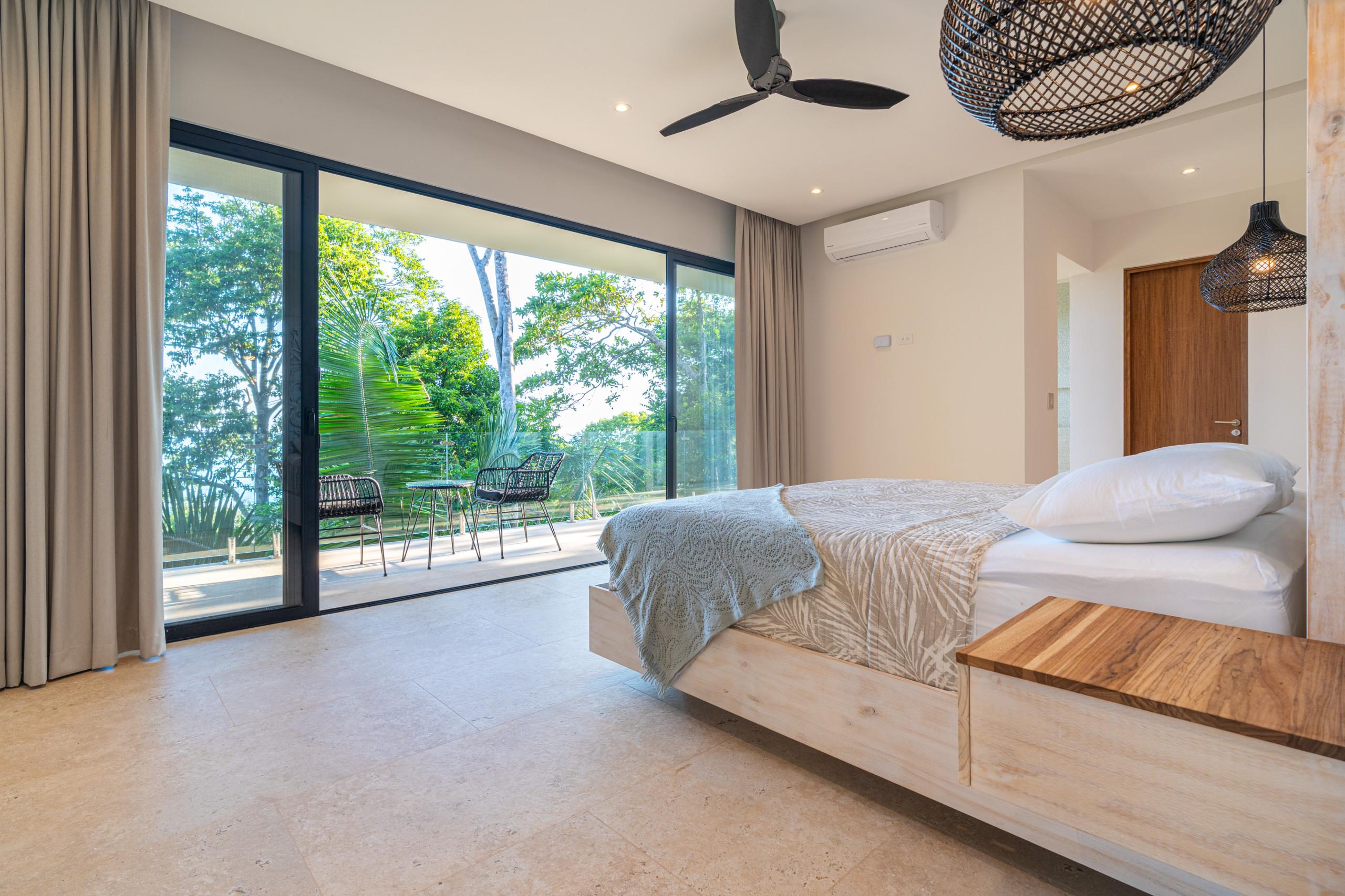 A bright bedroom features a king-size bed, ceiling fan, and air conditioning, with floor-to-ceiling glass doors opening to a private balcony and tropical jungle views.