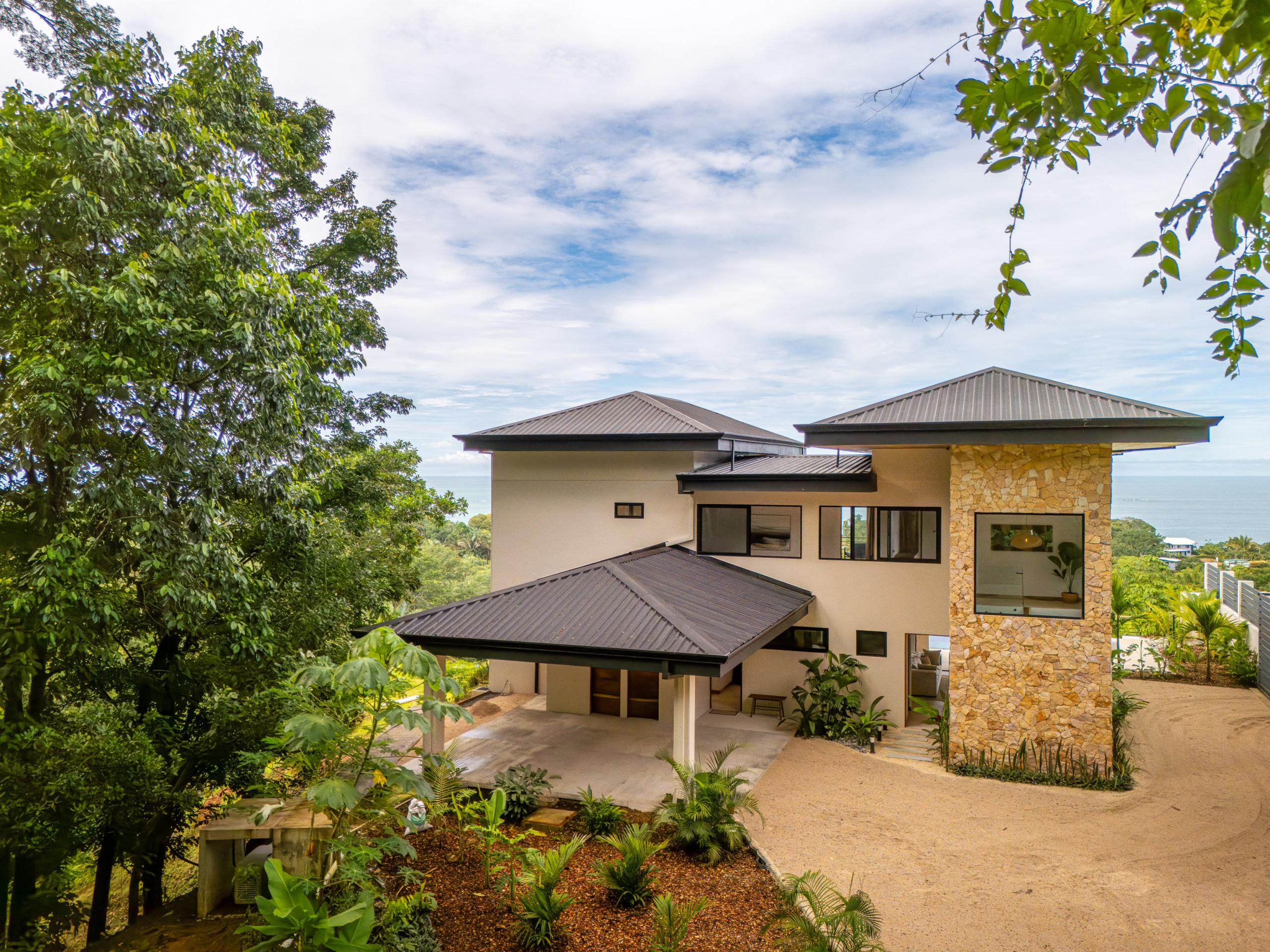 This contemporary multi-level villa features striking stone and stucco exterior walls with dark roofs, set amidst tropical trees and offering distant ocean views under an expansive sky.