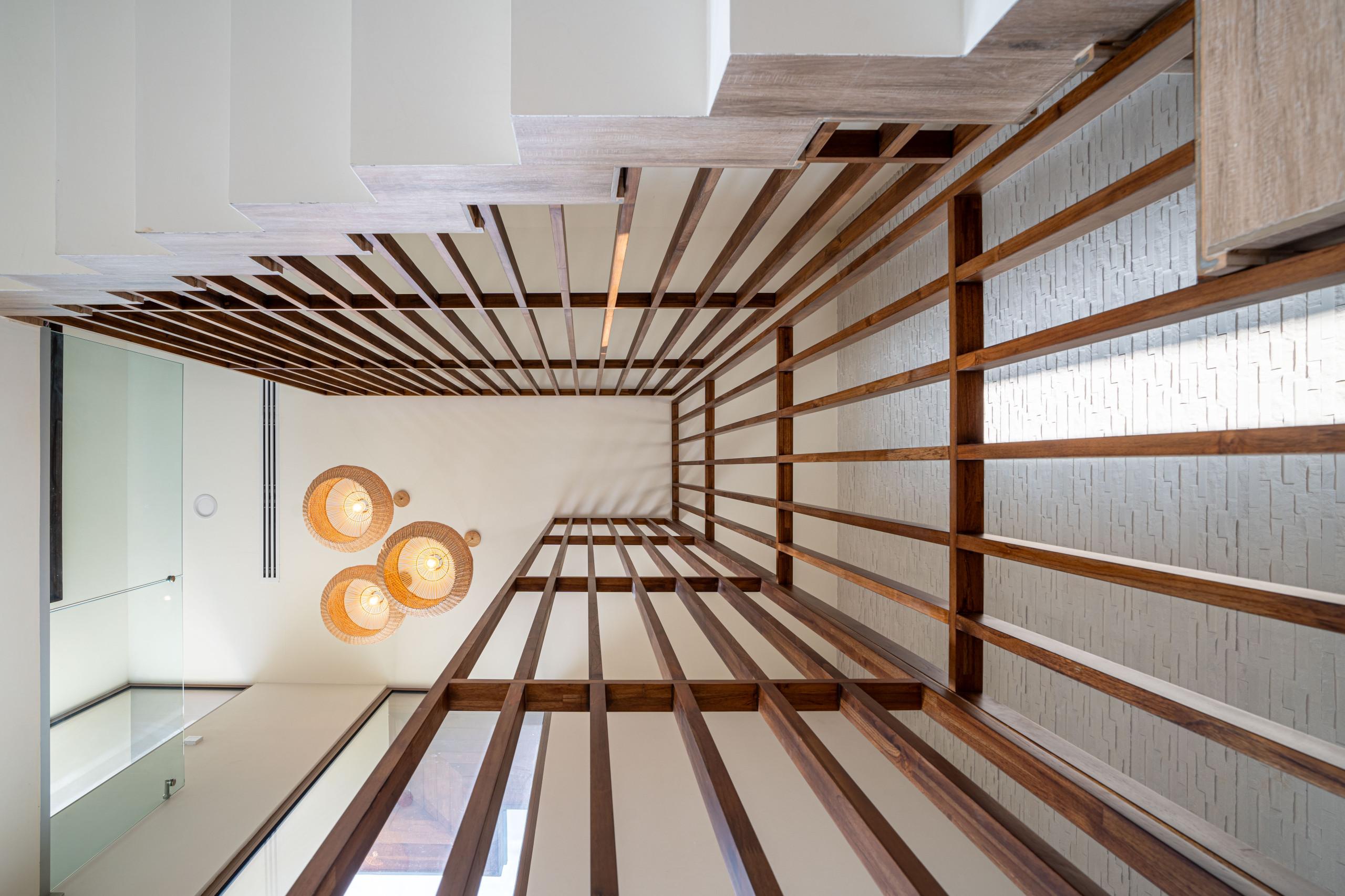 An upward view captures a multi-level interior with dark wooden ceiling slats, a white staircase and glass railing, three woven pendant lights, and a textured accent wall, emphasizing natural materials.