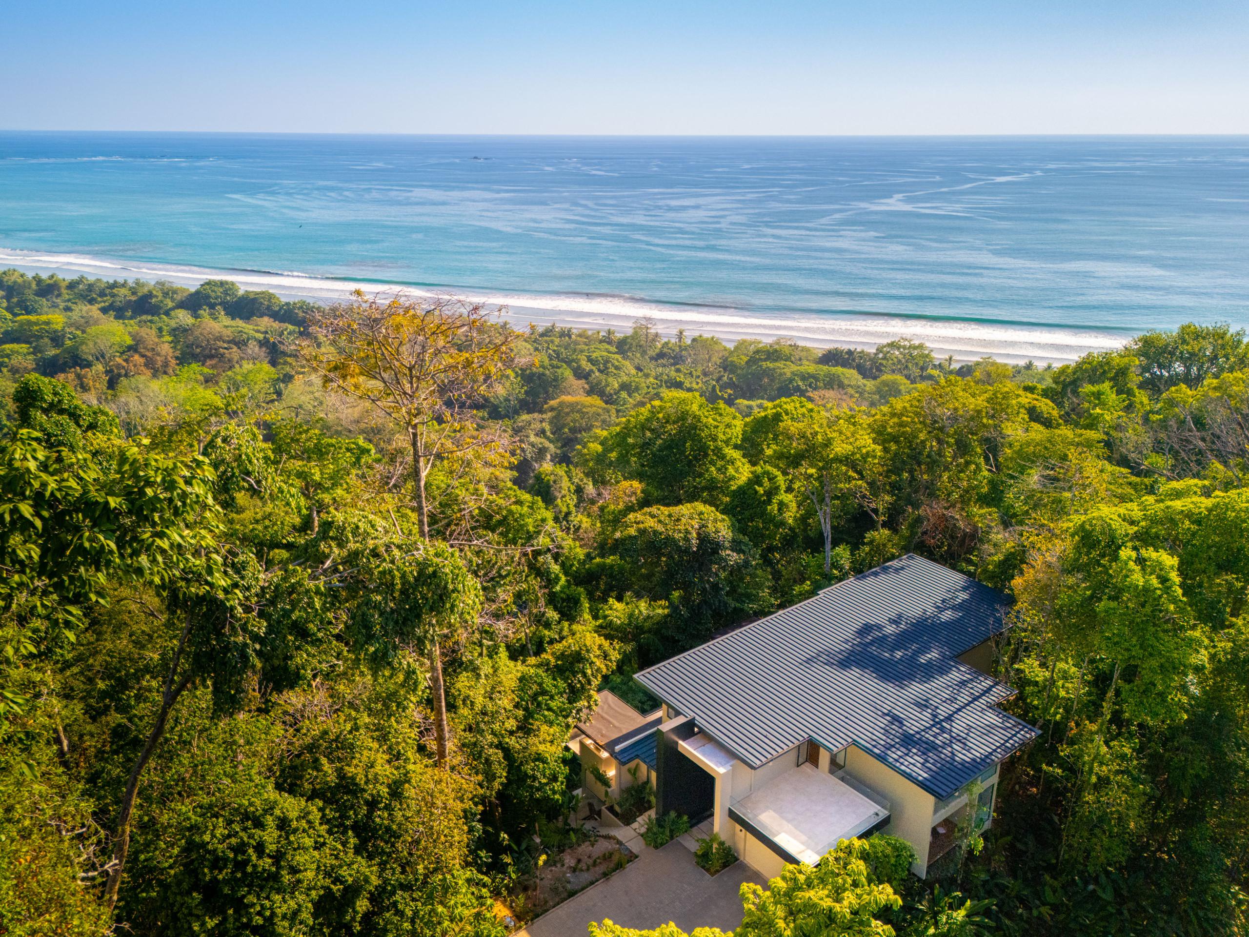 An aerial perspective reveals a modern villa nestled within a rich tropical forest, offering expansive views of the Pacific Ocean and the adjacent coastline.